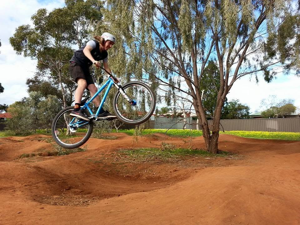 Rider in a helmet launching a blue BMX-style bike off a dirt roller under gum trees